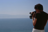 A figure with dark, short hair wearing a brown shirt and white pants looks into the viewfinder of a large videocamera. They appear to stand on a precipice above the ocean, and their camera is aimed at the water below them.