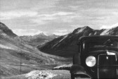 A grainy, black and white image of a vintage automobile driving on a winding, deserted road through a set of mountains. The car is in the foreground of the image, with part of its body cut off by the image's frame.