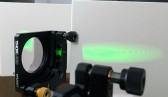 Detail view of an interferometer emitting a green light onto a white wall.
