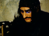 A figure wearing all black clothing, a black hood, heavy eyeshadow, and a fake mustache looks at the floor.