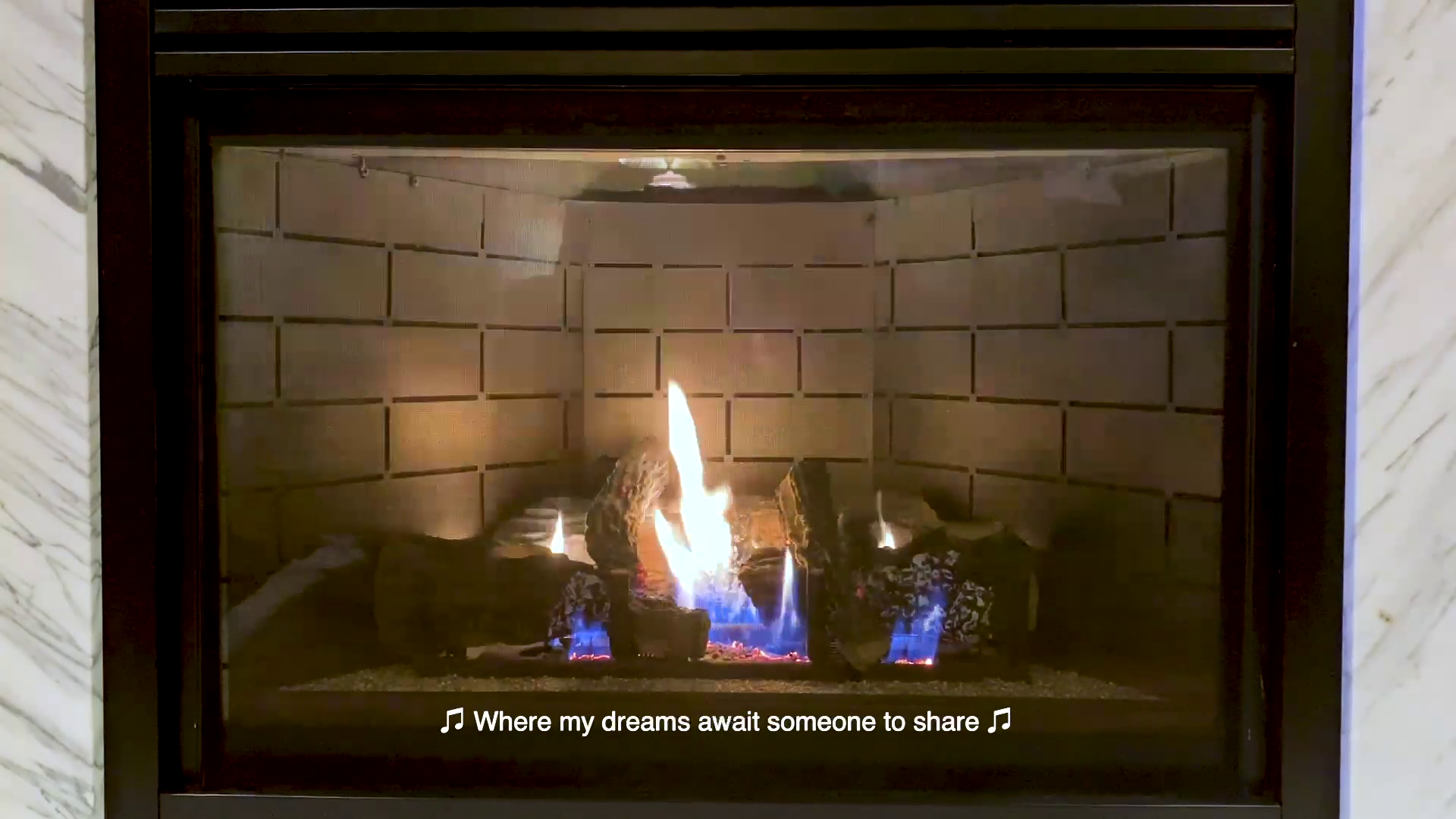 A gas fireplace turned on is framed with a strip of faux white marble on either side. The flames are yellow and white with glowing orange embers at their base. A yellow caption appears at the base of the fireplace with the following text in between musical notes: Where my dreams await someone to share.