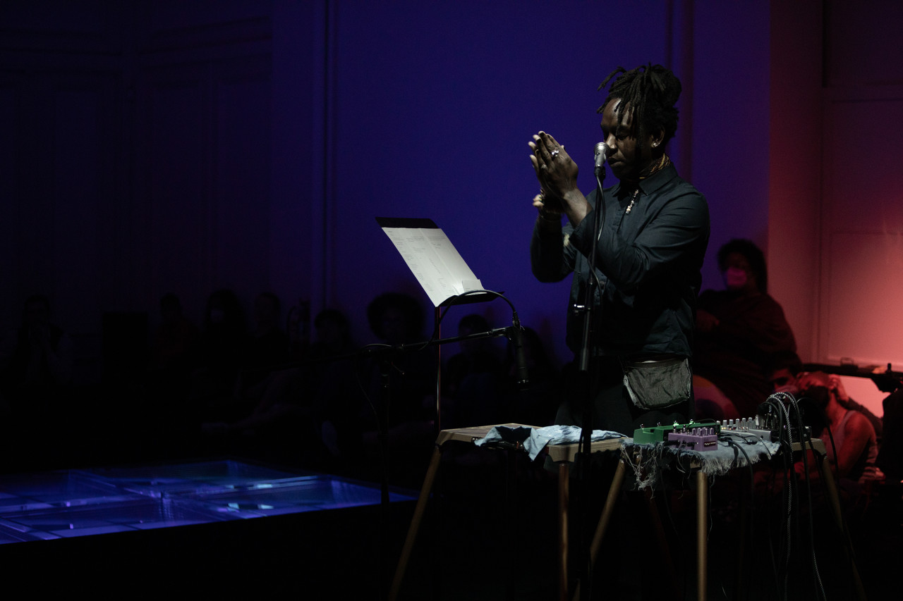 Photograph of a performer standing behind a small desk that holds audio equipment. The performer stands upright with their head down and their hands held together in front of their face. To the performer