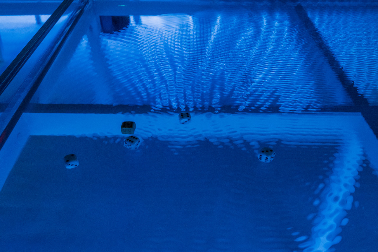 Five dice float at the top of a container of water, which is tinted blue. There are small ripples created across the surface of the water and the frame of structure containing the water is slightly visible through the ripple distortion.