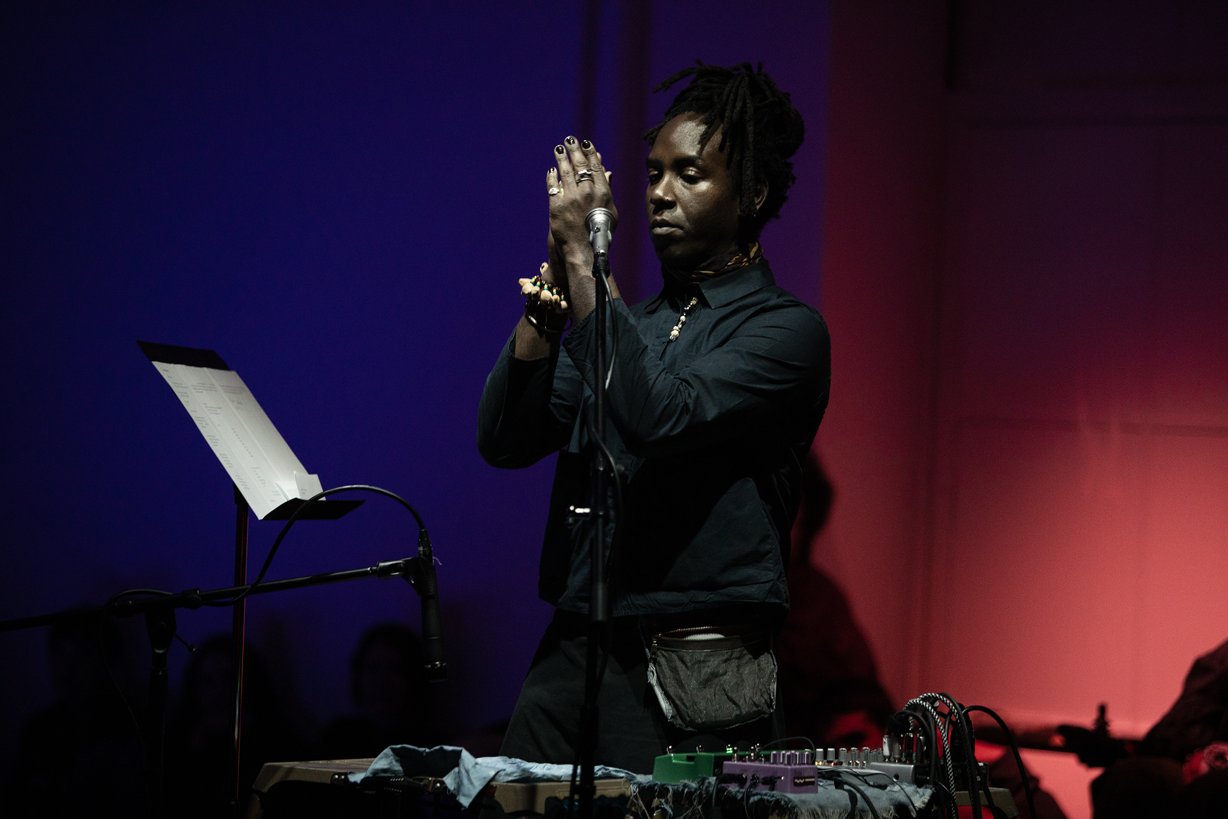 Photograph of a performer standing behind a small desk that holds audio equipment. The performer stands upright with their hands held together in front of their face. To the performer's right a music stand holds a score. The background is bathed in blue and red light. 