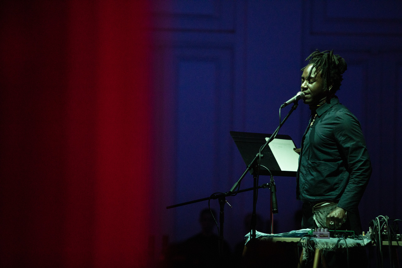 A performer closes their eyes as they sing into a mic pressed up to their mouth. They are holding a music stand with a score on it in their right hand, and adjusting audio equipment with their left hand. They are bathed in green light. On the left of the image a red-tinted column is blurred in the foreground. The background is dark purple.