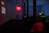 A group of people sit on the floor of a dimly-lit gallery. The audience is facing towards a wall on the left side of the image where a projection of live transcription text is projected. On the right side a performer bathed in green light holds their hands together as they stand infront of a mic. The audience member in the foreground is lit with red light, as well as an exit sign on the back wall. The rest of the gallery walls are blue and purple tinted.