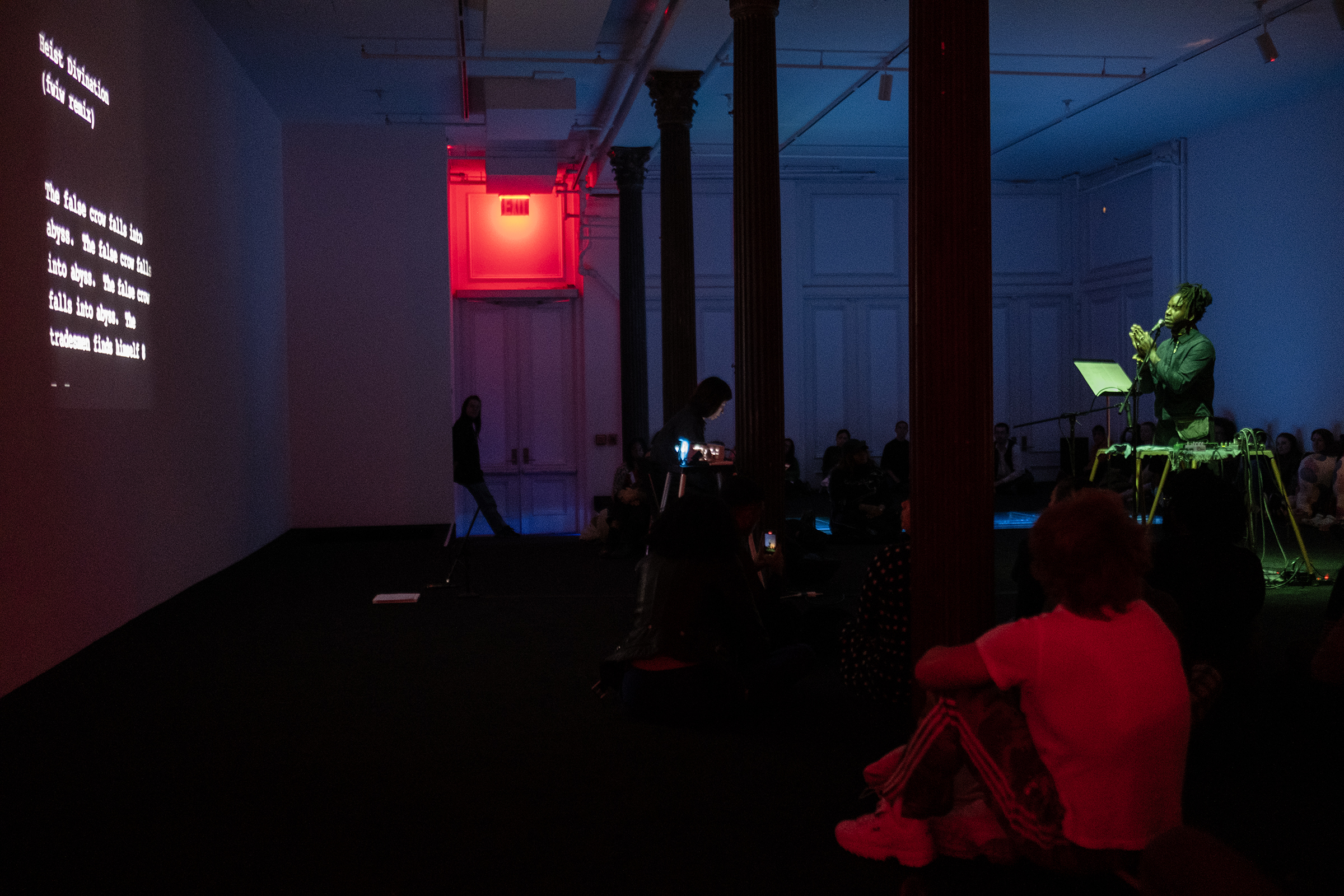 A group of people sit on the floor of a dimly-lit gallery. The audience is facing towards a wall on the left side of the image where a projection of live transcription text is projected. On the right side a performer bathed in green light holds their hands together as they stand infront of a mic. The audience member in the foreground is lit with red light, as well as an exit sign on the back wall. The rest of the gallery walls are blue and purple tinted.