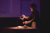 Photograph of a performer, standing next to a table covered in a black cloth. The background is bathed in purple light. A white spotlight shines on the table, highlighting the performer's hands as they pour liquid from one teacup into another teacup held below.