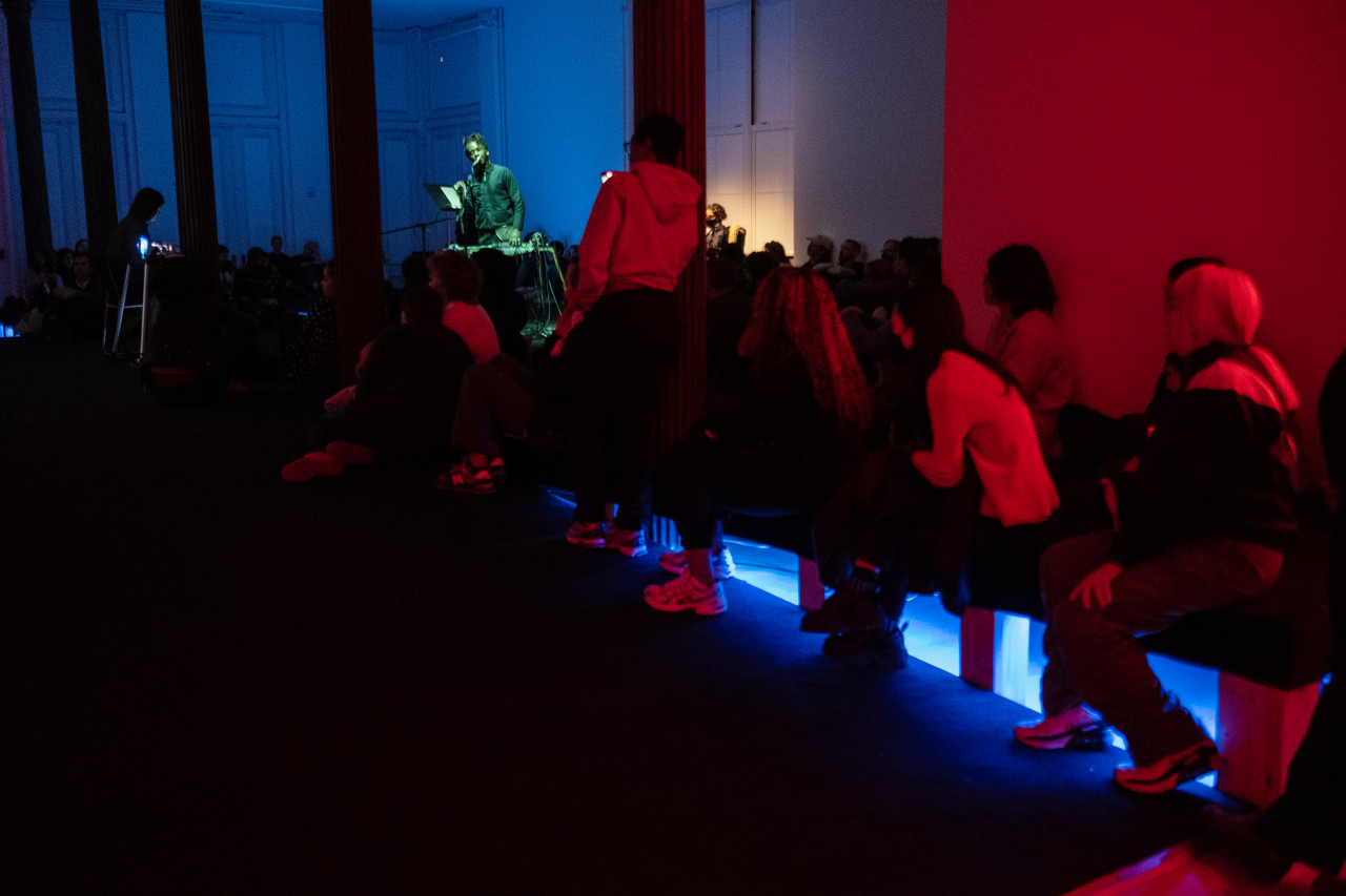 Audience members sit and stand alongside a ramp in a gallery. They are facing a performer who is framed in between columns to the left of the image. The audience members are bathed in red light, with a bright blue light coming from under a platform on the floor. The performer is lit with green light and the background is blue-tinted.