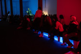 Audience members sit and stand alongside a ramp in a gallery. They are facing a performer who is framed in between columns to the left of the image. The audience members are bathed in red light, with a bright blue light coming from under a platform on the floor. The performer is lit with green light and the background is blue-tinted.