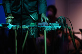 A performer's hand adjusts the dials of audio equipment which is placed on top of a small desk. Their hands are adorned with rings and bracelets, and the scene is bathed in green light. The background is blue and purple tinted, and there are blurred silhouettes of audience members behind the performer.