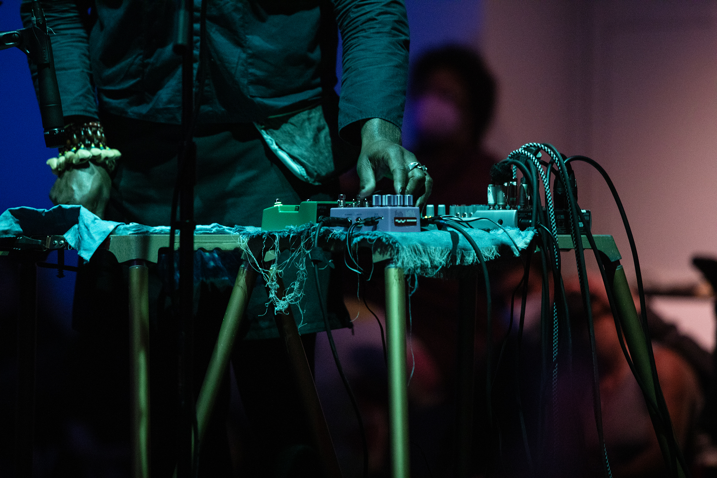 A performer's hand adjusts the dials of audio equipment which is placed on top of a small desk. Their hands are adorned with rings and bracelets, and the scene is bathed in green light. The background is blue and purple tinted, and there are blurred silhouettes of audience members behind the performer.