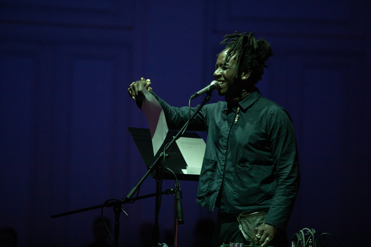 A performer stands close to a microphone with their right arm in the air holding a musical score. Their eyes are closed and pieces of their hair falls over their face. They are bathed in soft green light and the background is tinted dark purple.