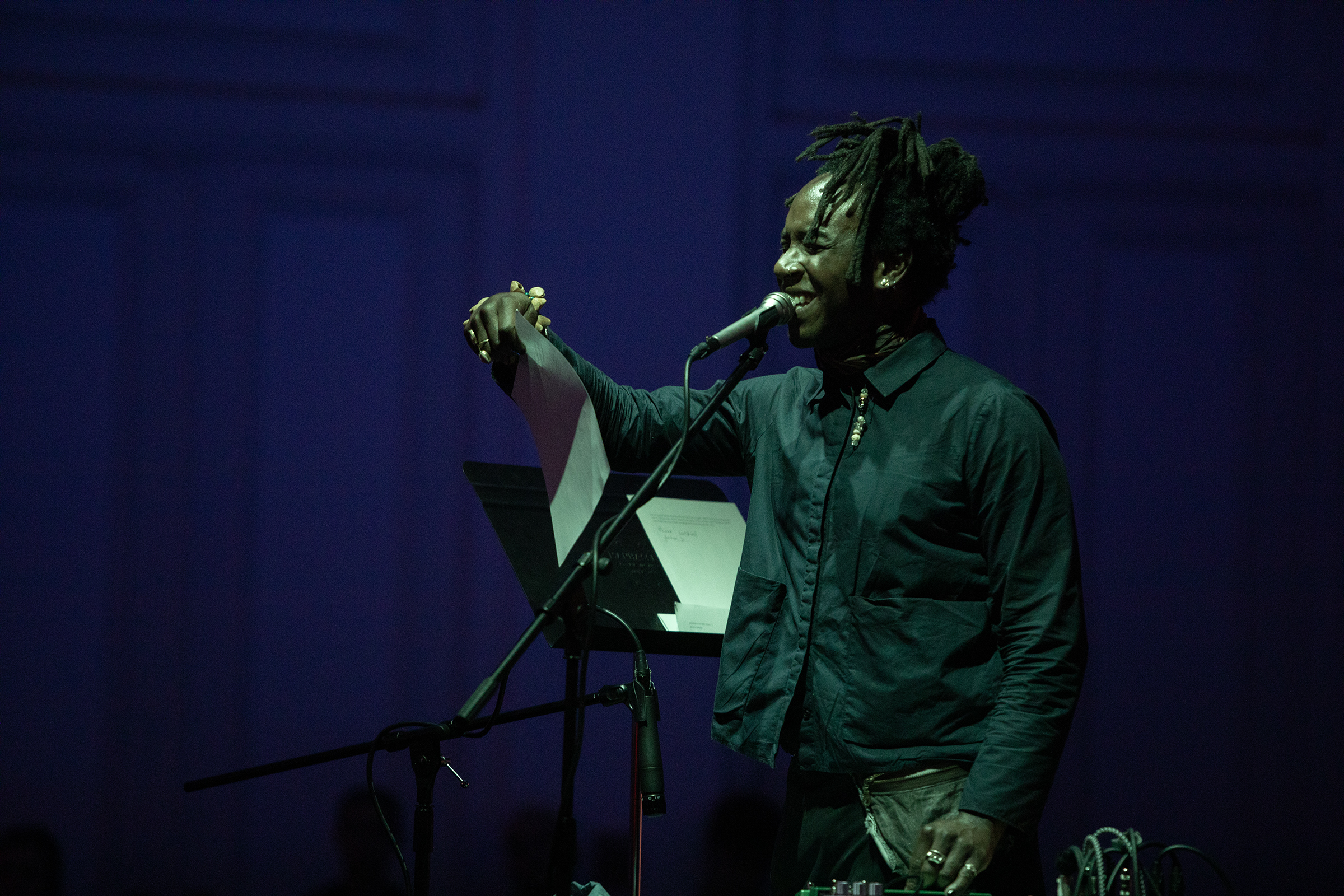 A performer stands close to a microphone with their right arm in the air holding a musical score. Their eyes are closed and pieces of their hair falls over their face. They are bathed in soft green light and the background is tinted dark purple.