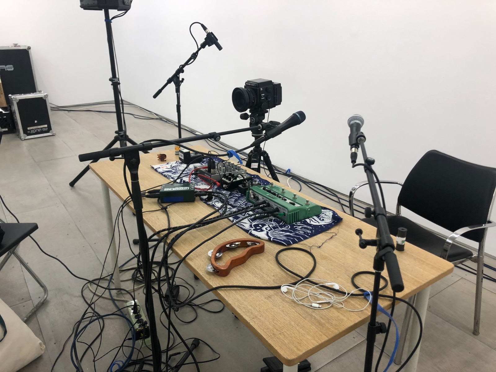 Photograph of a wooden table covered in a plethora of audio equipment, a tambourine, and wiring. Around the table are three microphone stands, that point at the empty space above a black chair behind the table. Next to the chair is a camera and tripod.