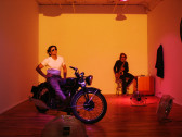 A person with sunglasses on a motorcycle. Behind him, a man in sunglasses sits on a stool playing a saxophone. There are three floor fans blowing toward the two people. The walls on either side are lit with a pink light, and the back wall with a yellow light.