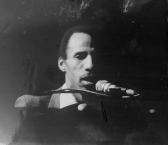 A black-and-white photograph of a figure sitting in front of a microphone. The left side of their face and body is in shadow.