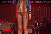 Close up of the torso and legs of a figure with long hair wearing nylon tights and a blue silk jacket on their bare chest. They are illuminated by red stage lights and appear to stand on a stage in front of a drum set and speakers.