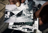 Close-up of two hands holding a series of black and white photographs, as if presenting them to the viewer. The top photograph depicts the roof of a small home that has caved in, battered by a recent storm. Debris and fallen branches are strewn around the home.
