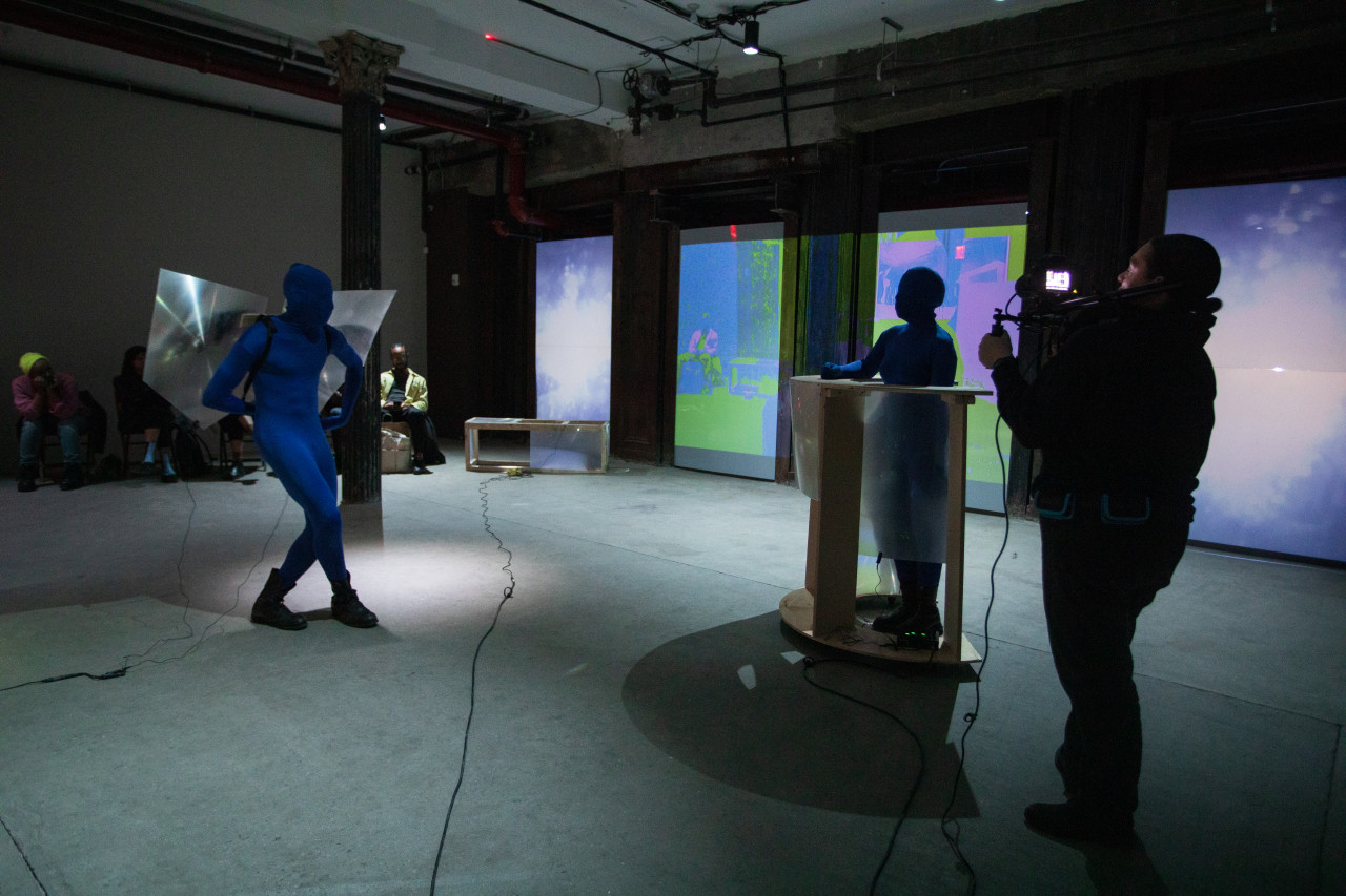 Three figures perform in a basement. On the left a figure in a blue morph suit wears a pair of wings made of fresnel lenses. On the right another figure in a blue morph suit stands behind a rolling table made of wood and fresnel lenses. Another figure to the far right takes video footage.