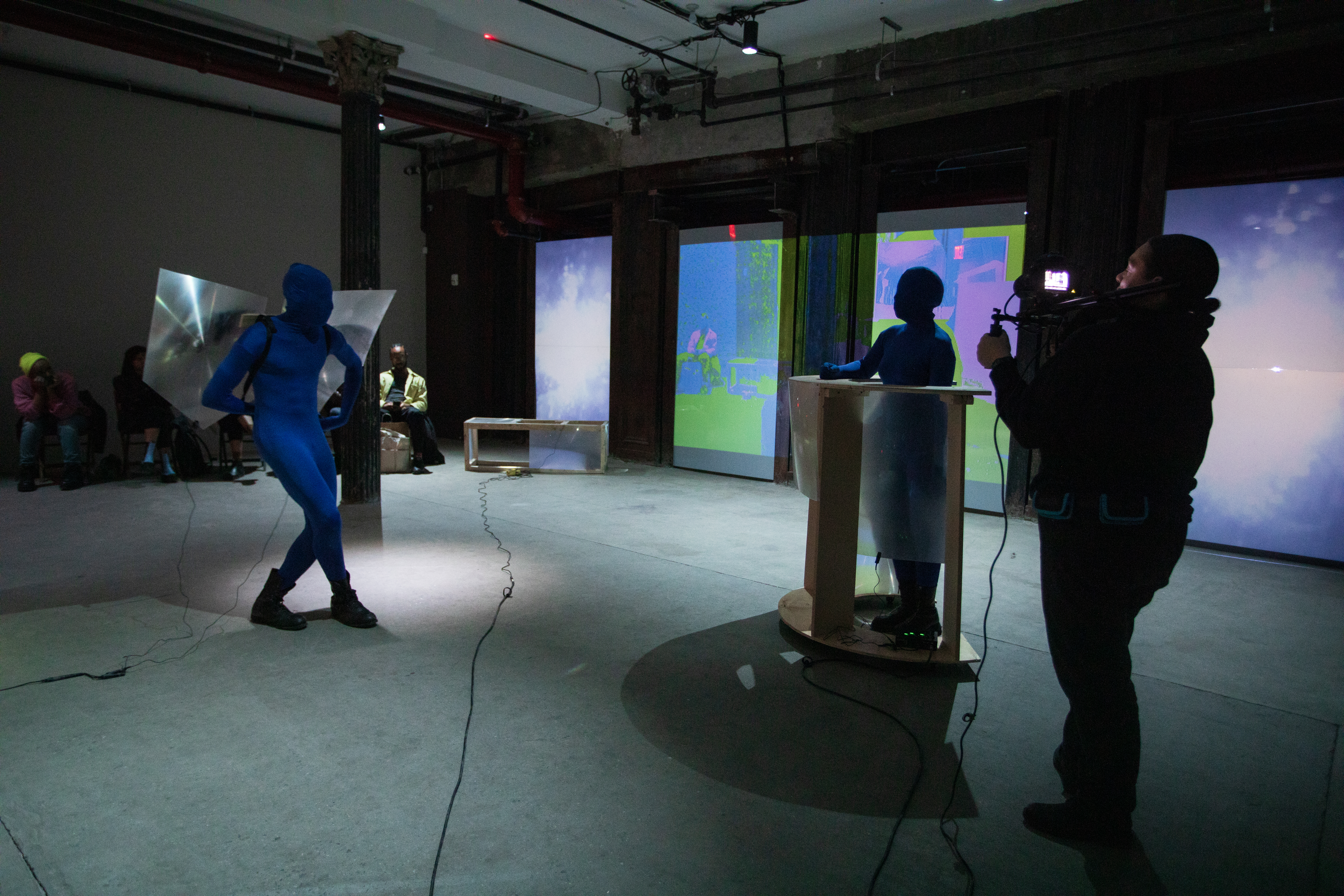 Three figures perform in a basement. On the left a figure in a blue morph suit wears a pair of wings made of fresnel lenses. On the right another figure in a blue morph suit stands behind a rolling table made of wood and fresnel lenses. Another figure to the far right takes video footage.