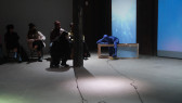 A figure in a blue morph suit leans over a wooden box placed on the floor, while another figure to the left films them. Blue light is projected onto the back wall to the right.