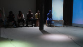 Audience members are seated along a back wall, with a figure in a blue morph suit seated on a rectangular wooden frame alongside them. Blue light is projected onto a screen in the background.