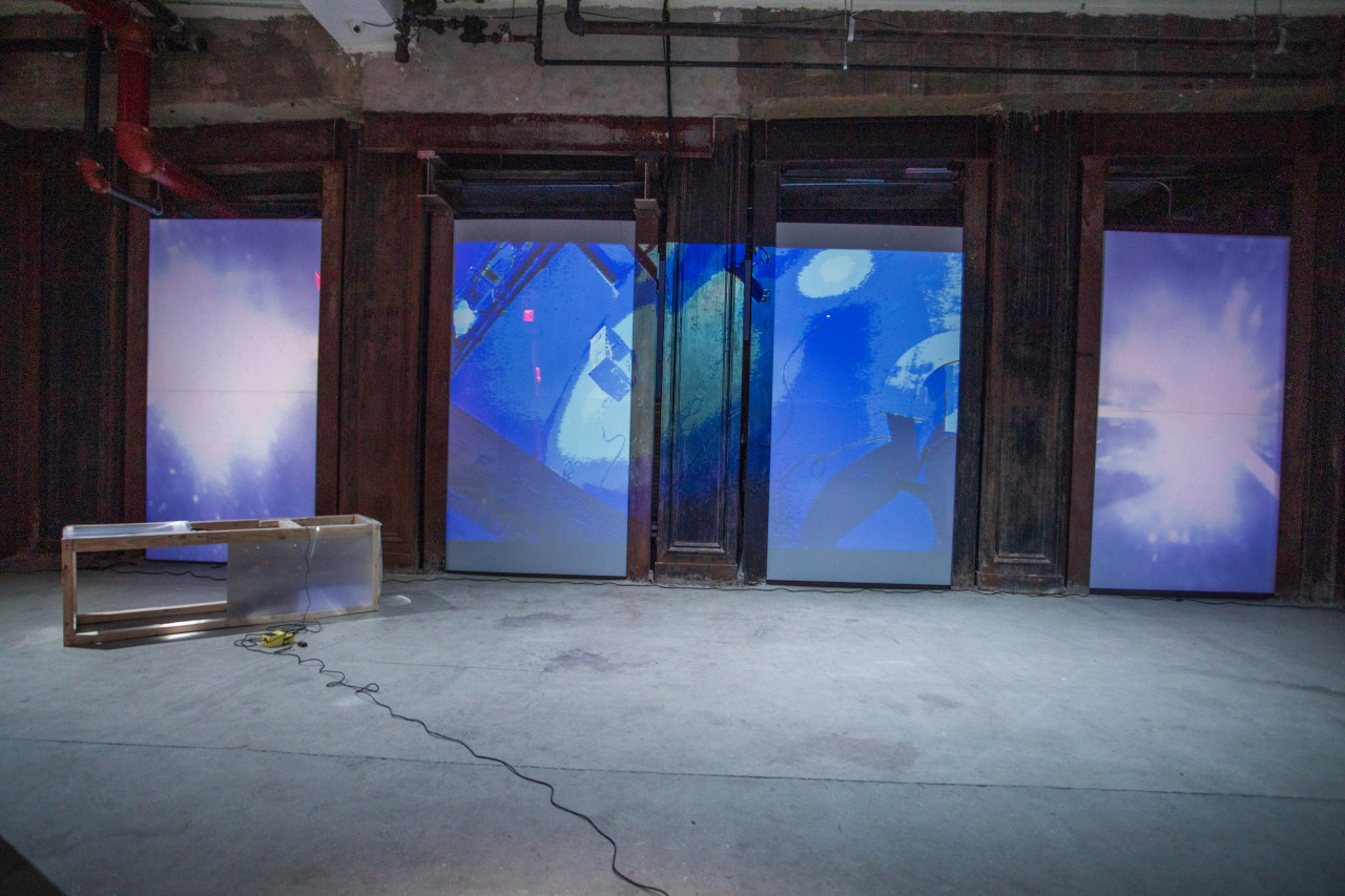 Image of a basement with a rectangular shaped sculpture made of a wooden frame with fresnel lenses placed along the top and sides. Along the back wall a blue projected image of live CCTV footage of the basement is displayed.