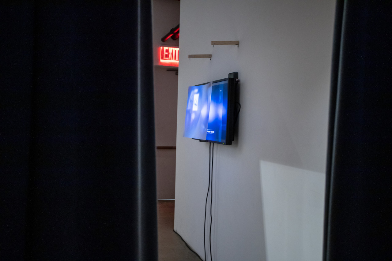 Image taken between two curtains of a TV affixed to a wall on the right, displaying blue-tinted video footage of the performance with a fresnel lens hanging in front of the TV display.