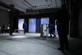 Three figures perform in a basement. Two figures in blue morph suits act out a scene together. The figure on the right scratches a fresnel lens attached to a wooden rolling table while the left figure walks toward the table. A third figure takes video footage of the scene.
