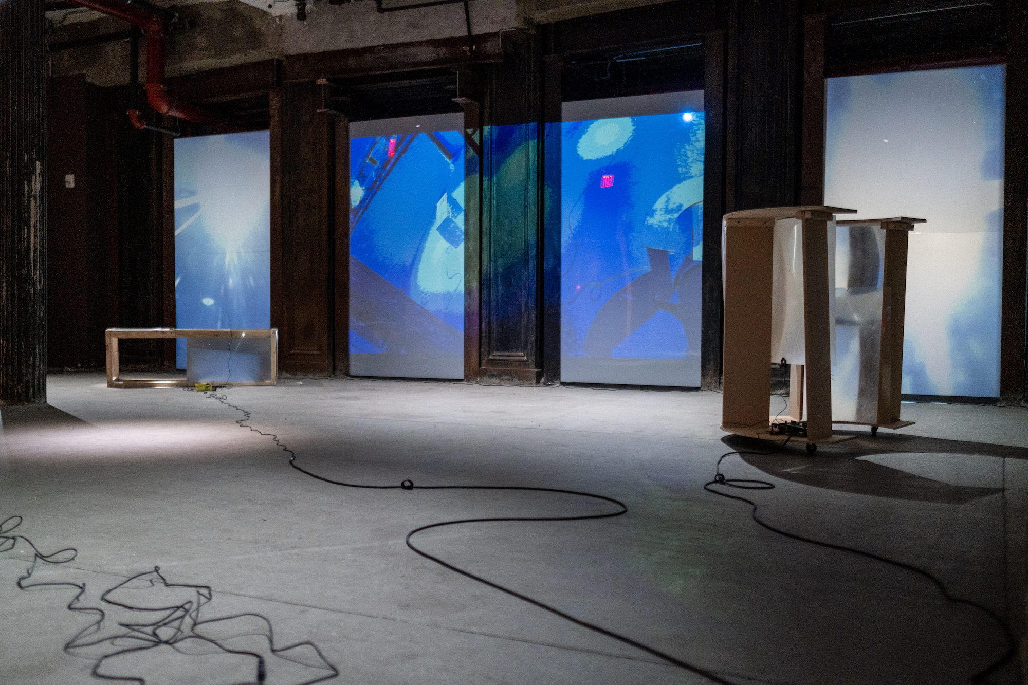 Image of a basement with two sculptures made of wood and fresnel lenses placed on either side of the image. Along the back wall a blue projected image of live CCTV footage of the basement is displayed.