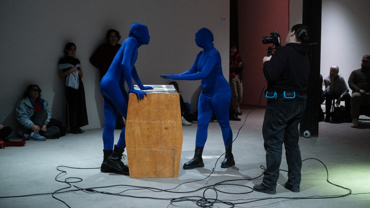 Two figures in blue morph suits engage in an argument, standing on either side of a wooden table with a fresnel lens as the tabletop. A third figure to the right films their encounter, while audience members watch in the background.