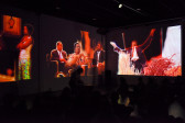 A crowd is pictured in front of a performer. Behind the performer are three large colorful projections.