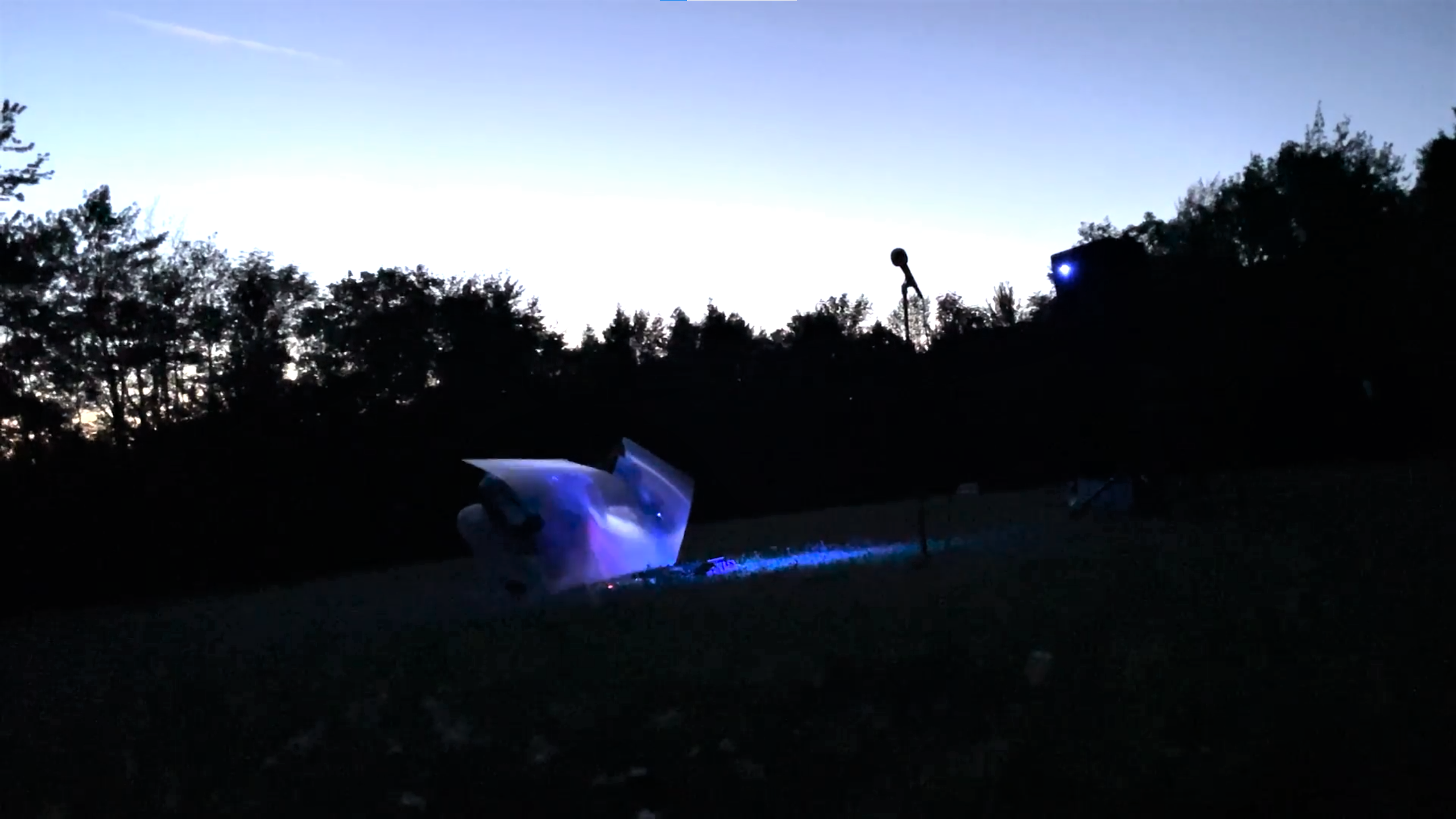 A microphone and lights are set up in a field, with trees along the horizon line.