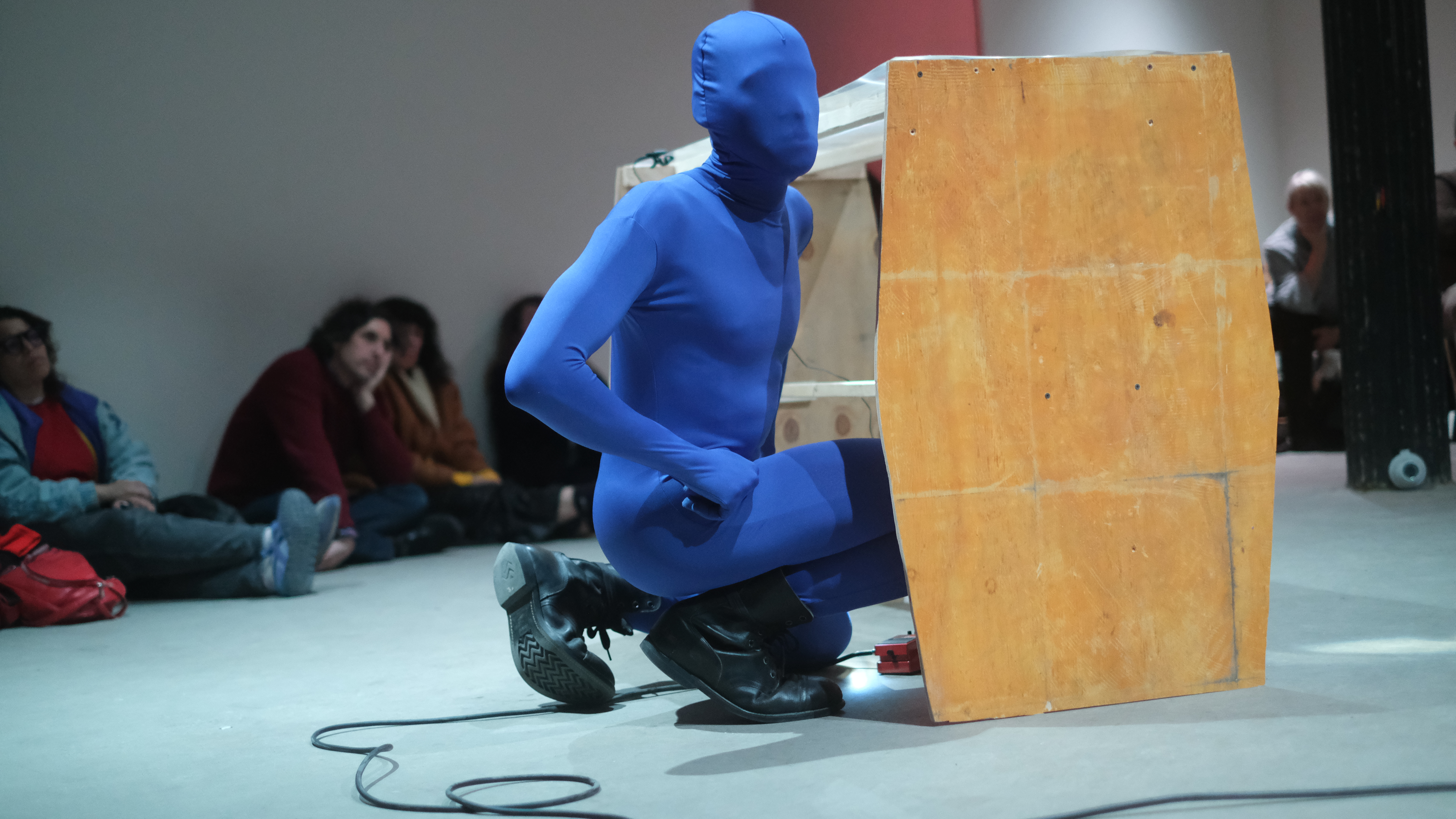 A figure in a blue morph suit kneels down next to a wooden table and looks off to the right. Audience members sit along the floor in the background.