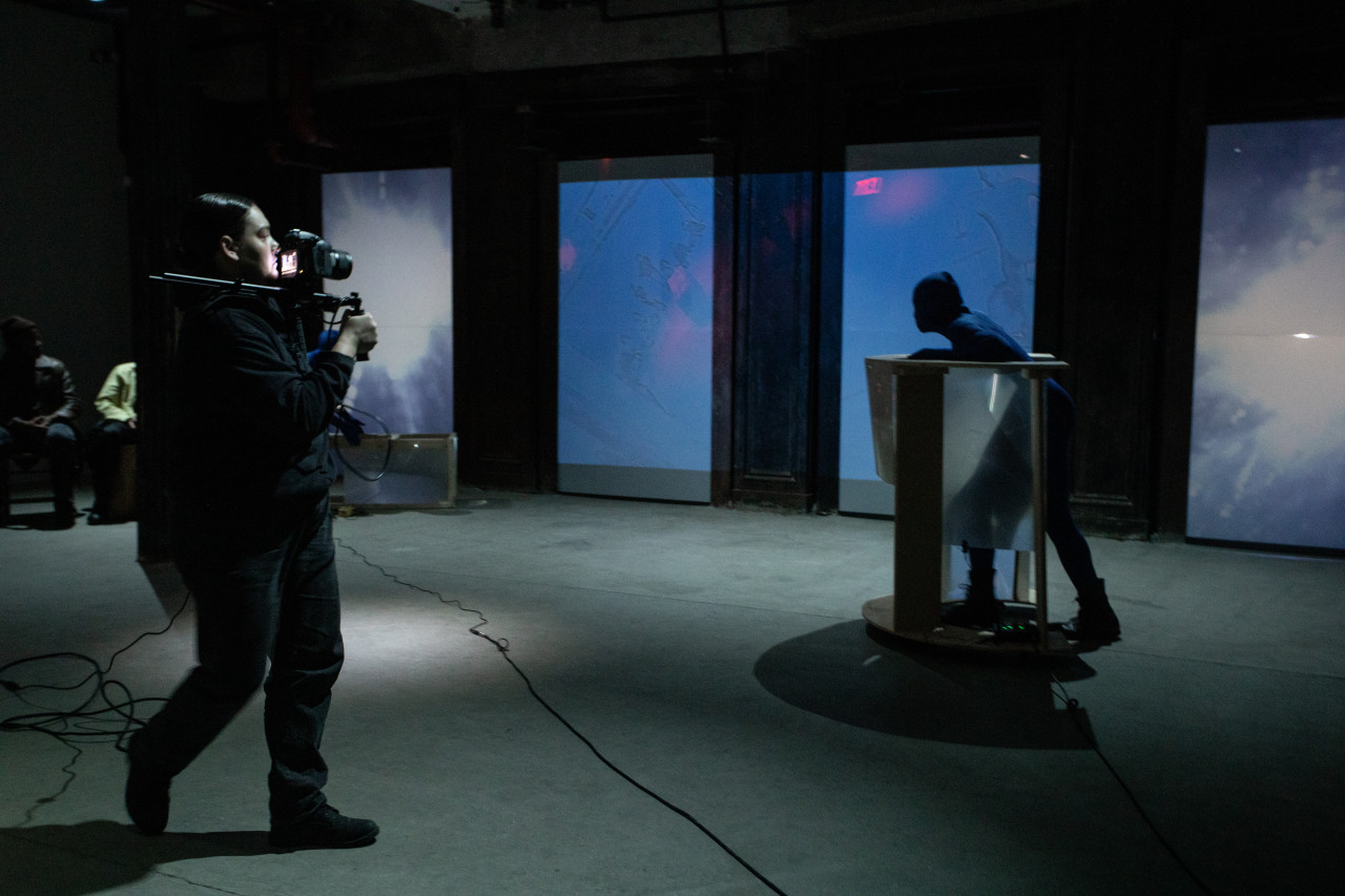 Two figures perform in a basement. On the left a figure with a video camera and shoulder rig takes footage of another figure on the right, dressed in a blue morph suit, and pushing a semicircular rolling table made of wood and fresnel lenses. Along the back wall blue and purple tinted live CCTV video footage is projected.