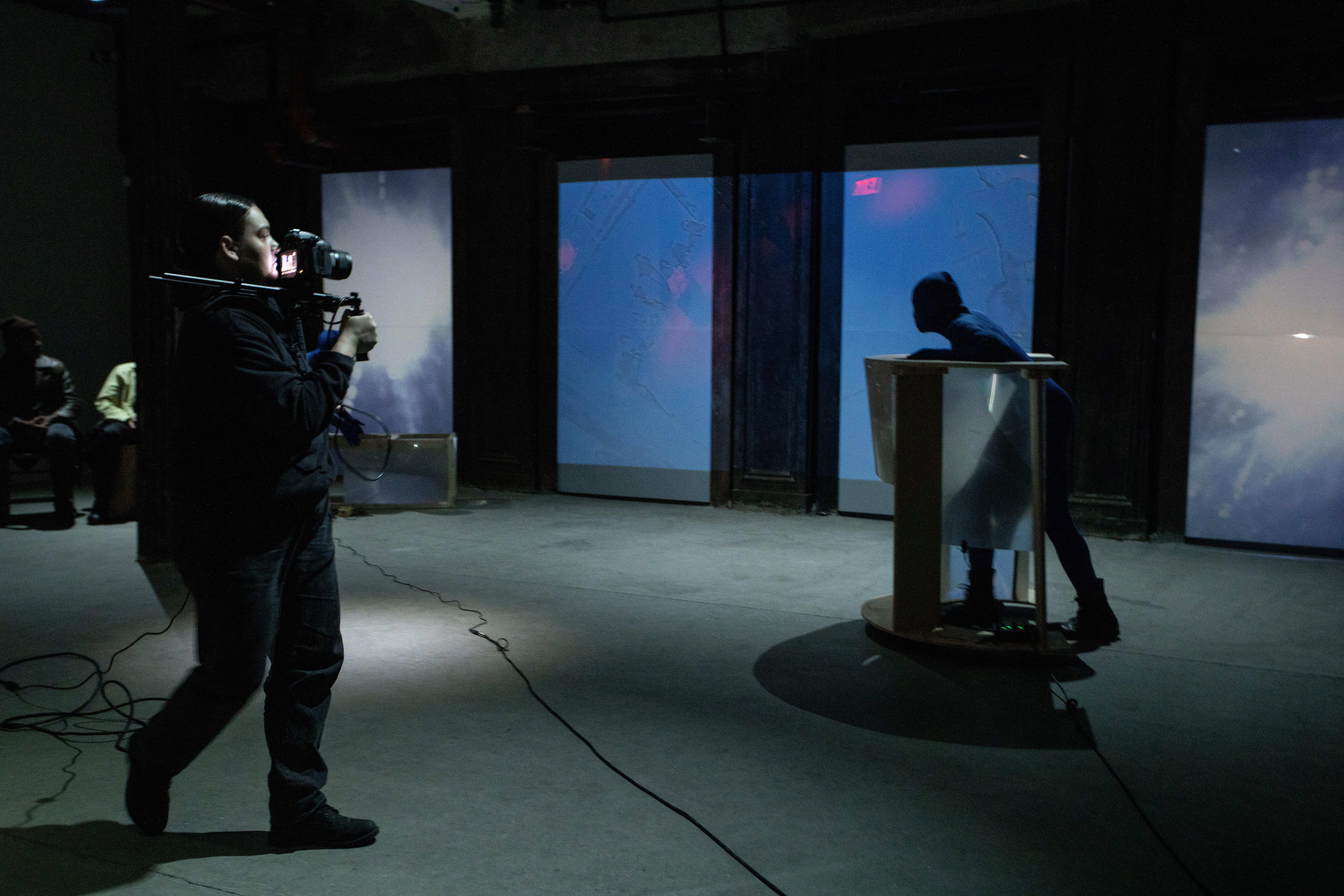 Two figures perform in a basement. On the left a figure with a video camera and shoulder rig takes footage of another figure on the right, dressed in a blue morph suit, and pushing a semicircular rolling table made of wood and fresnel lenses. Along the back wall blue and purple tinted live CCTV video footage is projected.
