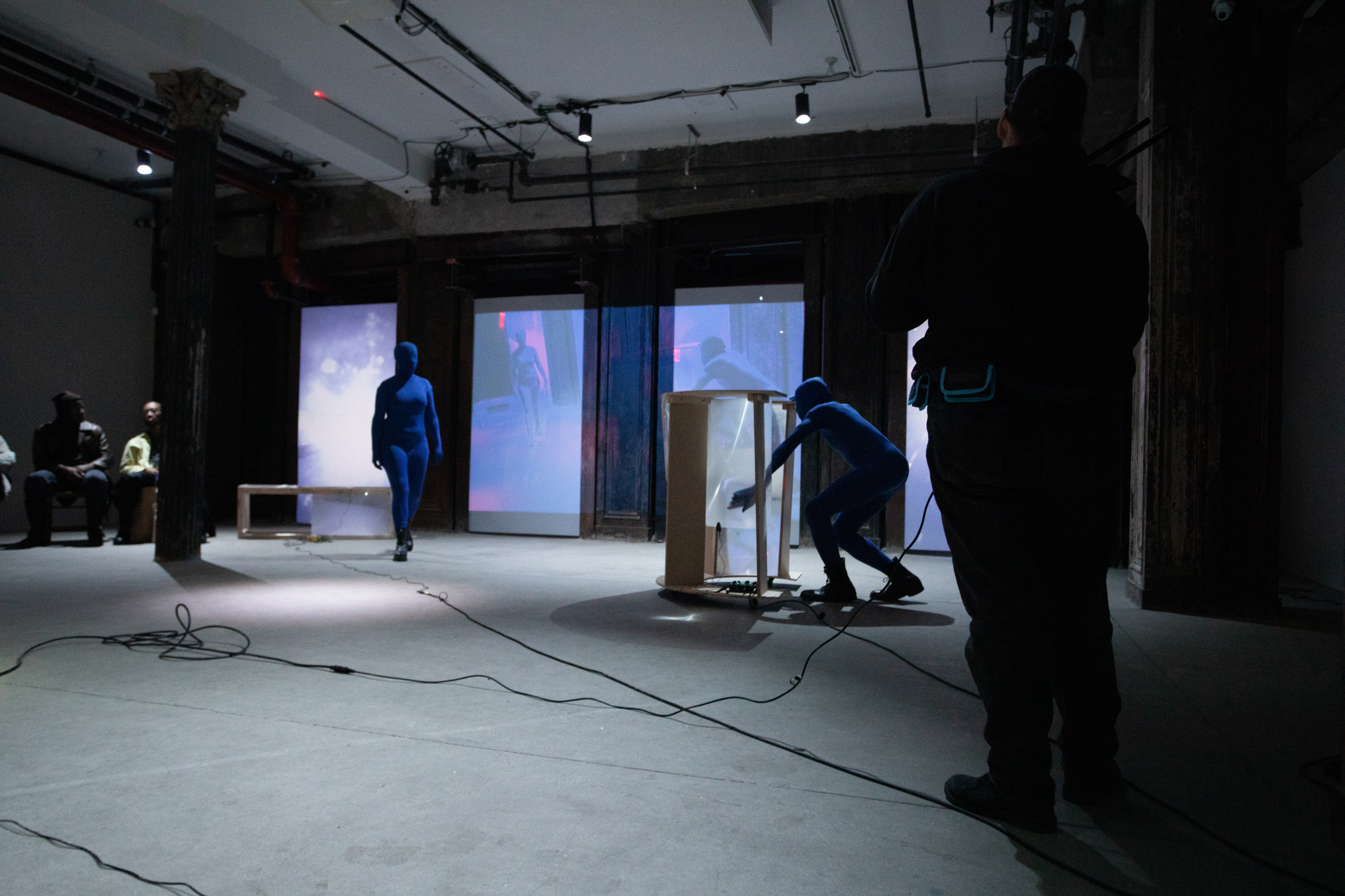 Three figures perform in a basement. Two figures in blue morph suits act out a scene together. The figure on the right scratches a fresnel lens attached to a wooden rolling table while the left figure walks toward the table. A third figure takes video footage of the scene.