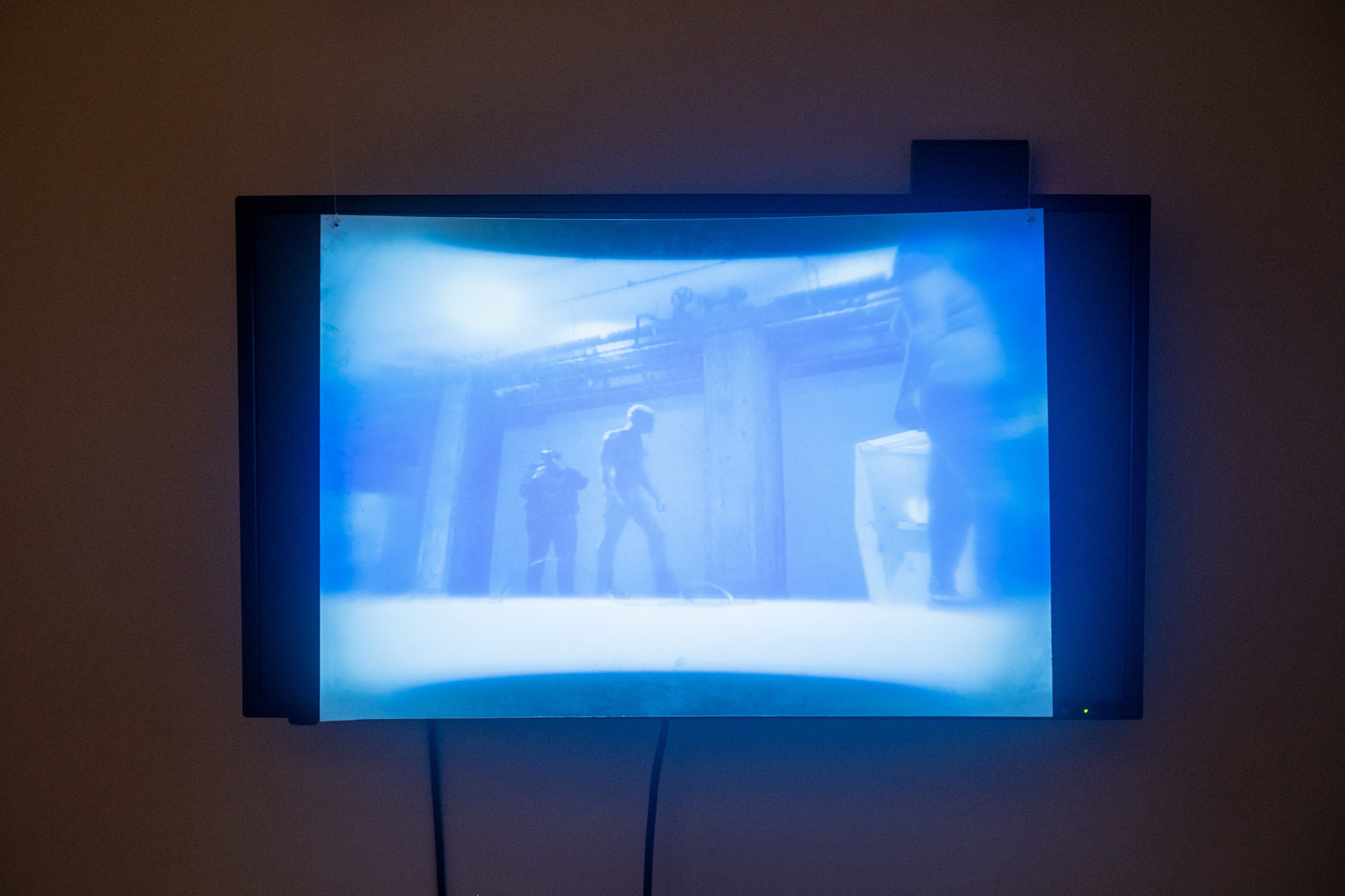 Image of a TV screen playing blue-tinted video footage of the performance with a fresnel lens placed in front of the screen, distorting the image.