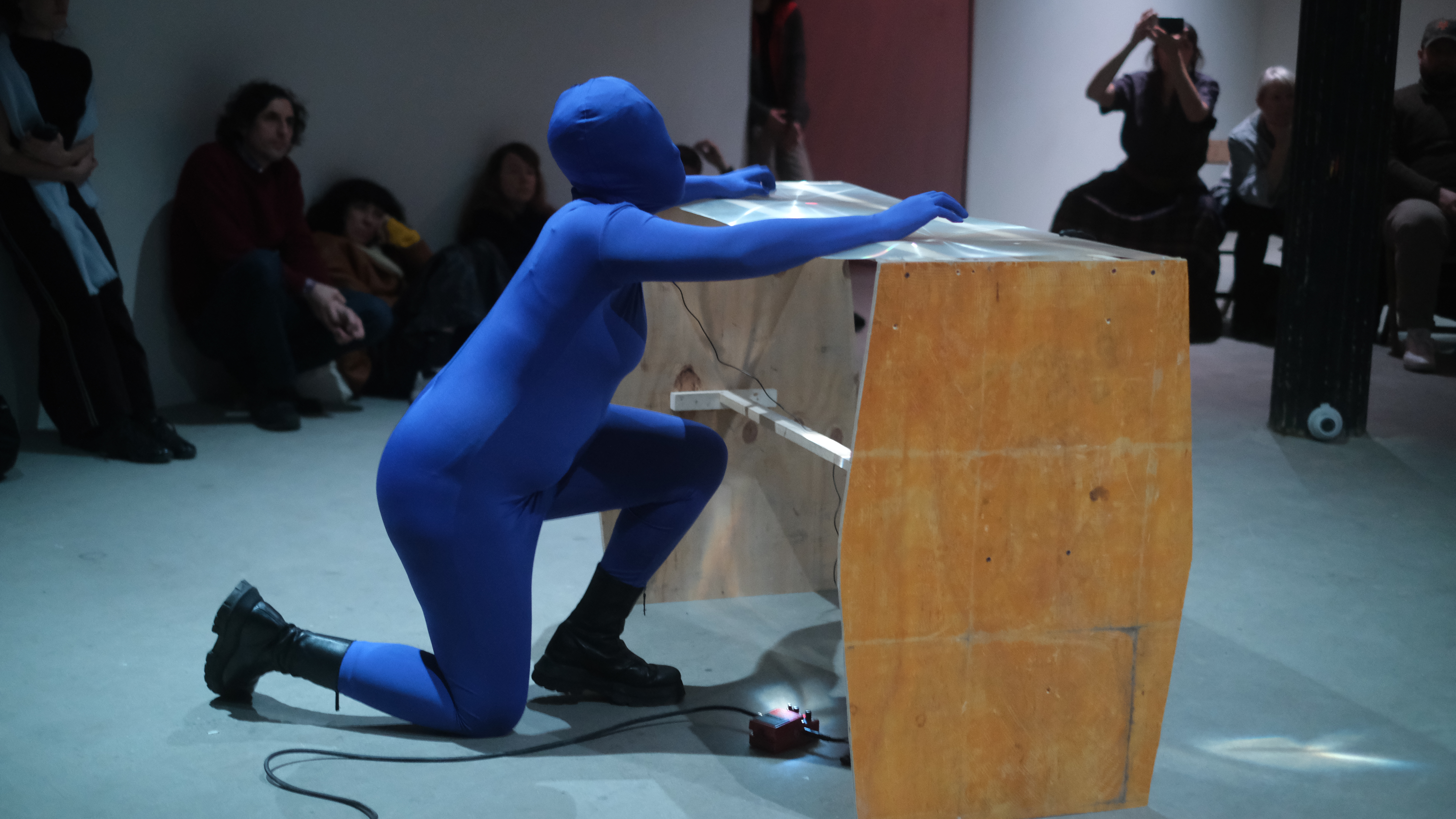 A figure in a blue morph suit kneels down on one knee, placing their hands on top of a table made of wood and fresnel lenses while audience members watch in the background.