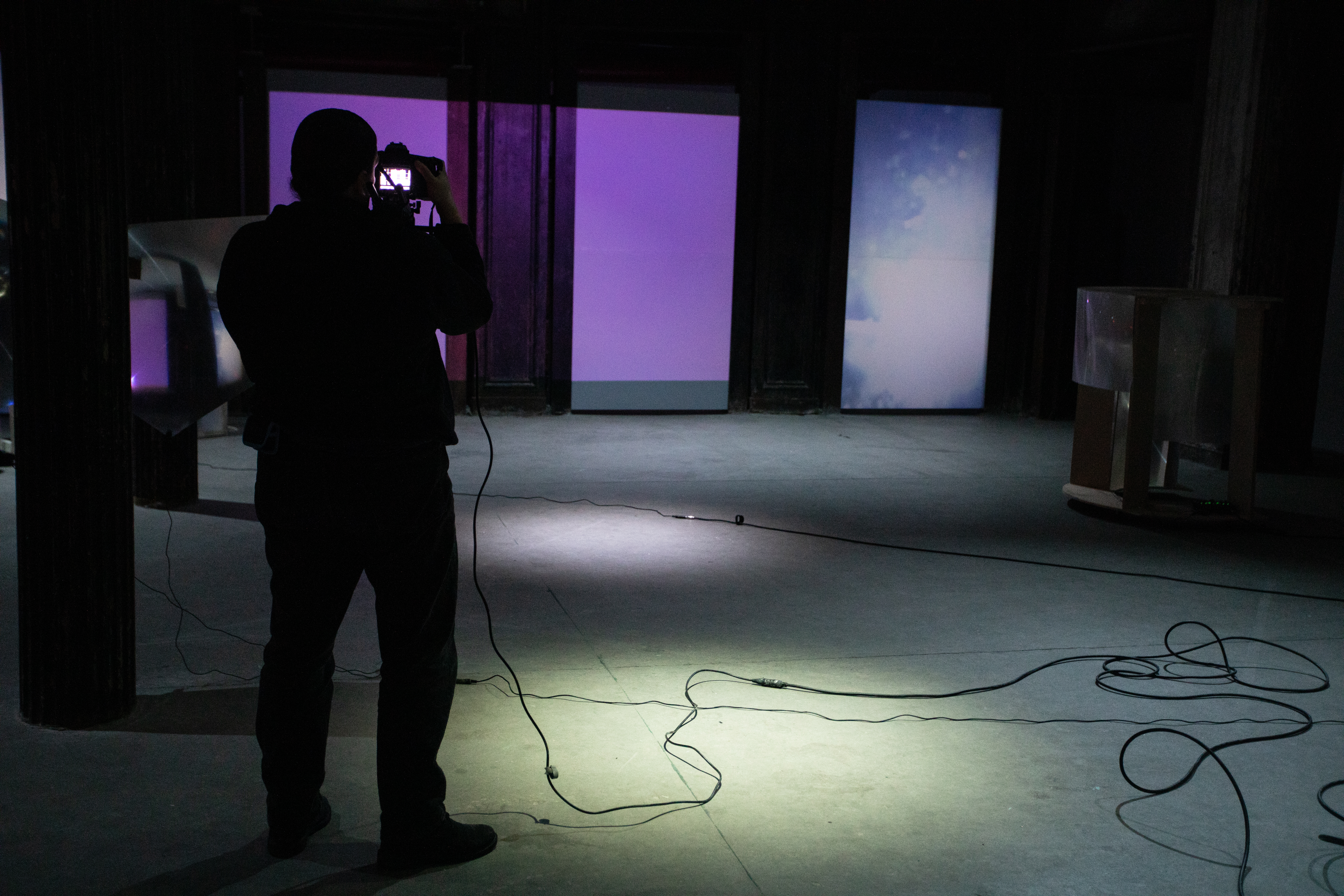 Image of a dimly lit basement, a figure holding a video camera with a rig on their shoulder takes footage of the back wall, which has blue and purple light projected onto it.