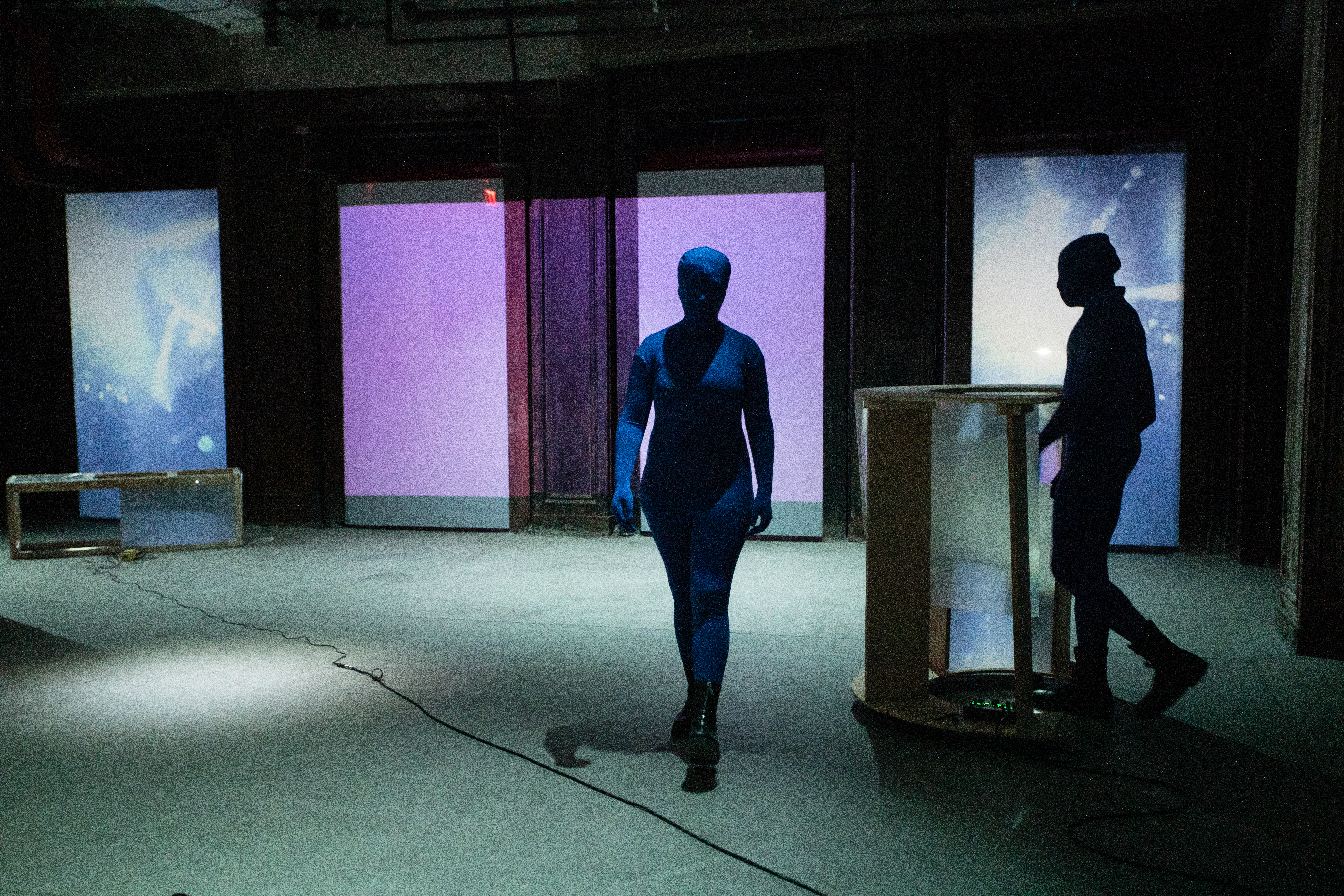 Two figures dressed in blue morph suits walk around a basement space. One figure on the right stands behind a semi-circular rolling table made of wood and fresnel lenses. Along the back wall blue and purple live CCTV video footage is projected.