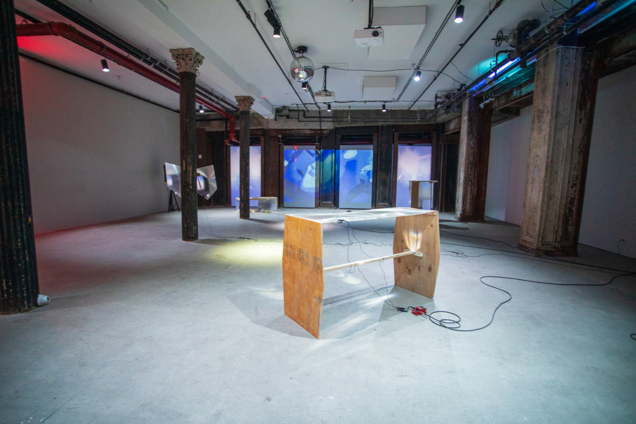Image of a basement with various sculptures made of wood and fresnel lenses placed around the space. Contact microphones are attached to the fresnel lenses and audio wires wrap around the floor. Along the back wall a blue projected image of live CCTV footage of the basement is displayed.