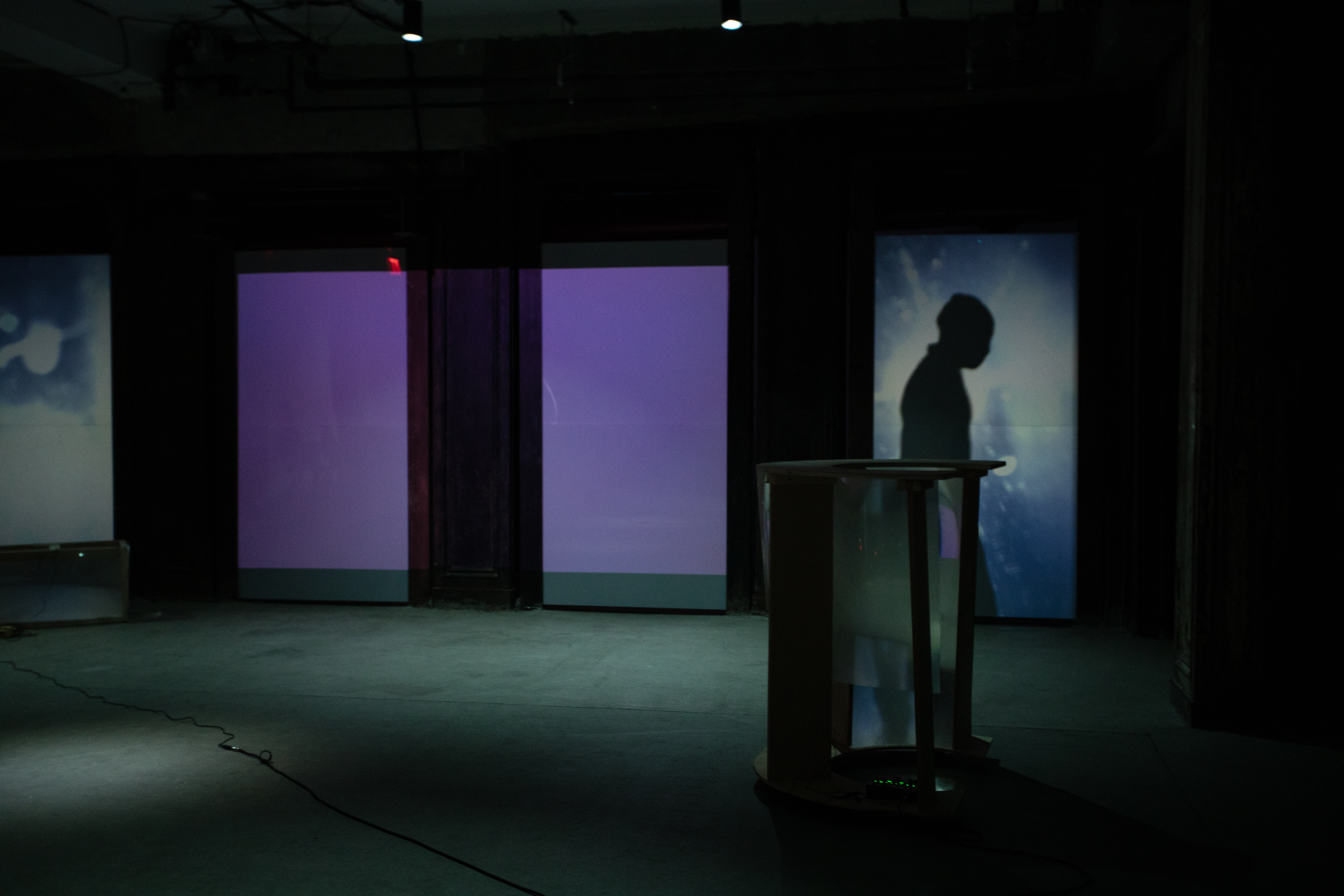 Image of a dimly lit basement. Along the back wall, on the right, a shadow of a figure in profile is cast onto a blue and purple projected screen.