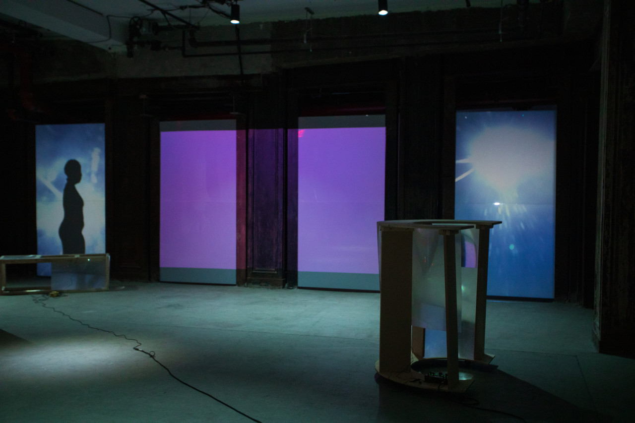 Image of a dimly lit basement. Along the back wall, on the left, a shadow of a figure in profile is cast onto a blue and purple projected screen.