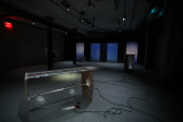 Image of a dimly-lit basement with various sculptures made of wood and fresnel lenses placed around the space. Contact microphones are attached to the fresnel lenses and audio wires wrap around the floor. Along the back wall a blue projected image of live CCTV footage of the basement is displayed.