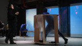 A figure in a blue morph suit leans their arms on top of a rolling table made of wood and fresnel lenses, pushing it toward another figure that stands in front of them filming them. In the background a third figure in a blue morph suit sits and leans down on top of a rectangular wooden frame. Blue footage is projected onto the back wall.