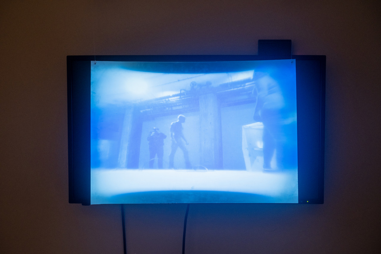 Image of a TV screen playing blue-tinted video footage of the performance with a fresnel lens placed in front of the screen, distorting the image.