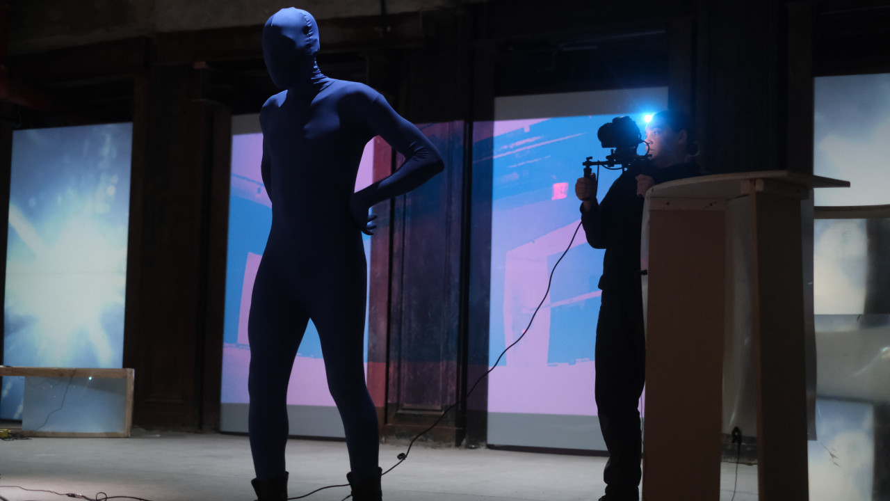 A figure in a blue morph suit stands, their hands on their waist, and looks to the left. A figure behind them films them. Blue video footage is projected onto the back wall.