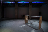 Image of a basement with a table made of wood and a fresnel lens tabletop on the right side of the space. In the back are audio wires and a speaker in the corner. Along the top of the image the basement windows are tinted blue.